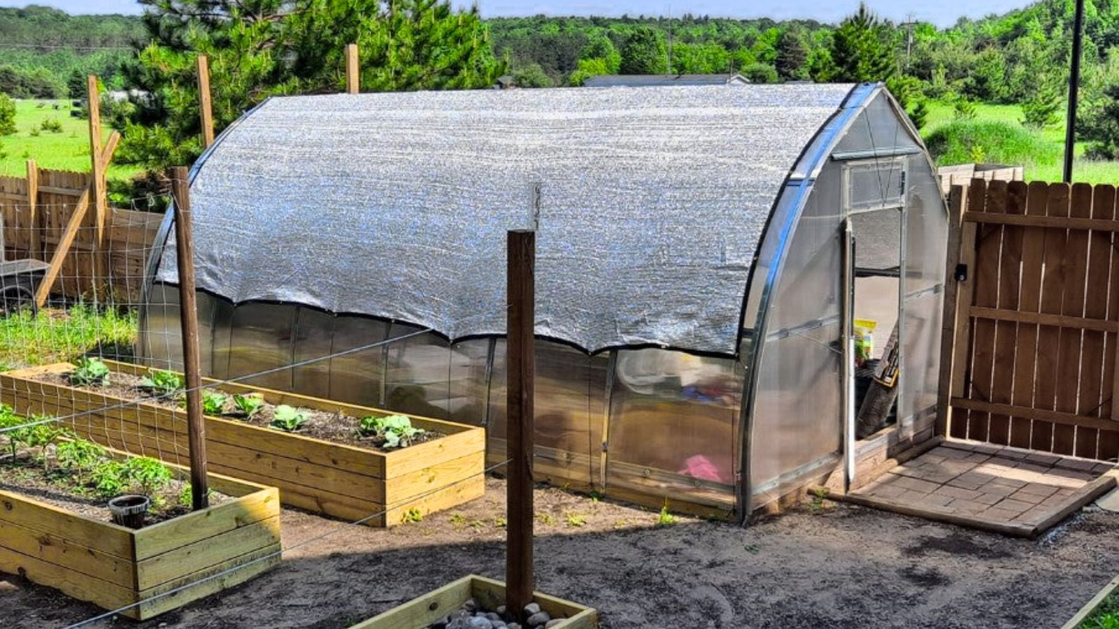 A greenhouse covered with a tarp, surrounded by a wooden fence, set in a garden environment.