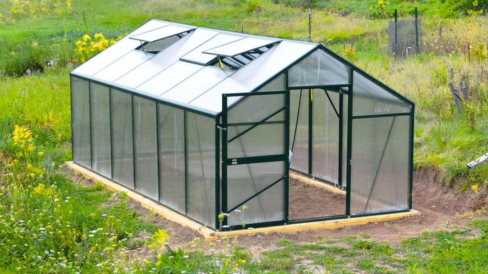 A greenhouse stands in a grassy field adorned with colorful flowers under a clear blue sky.