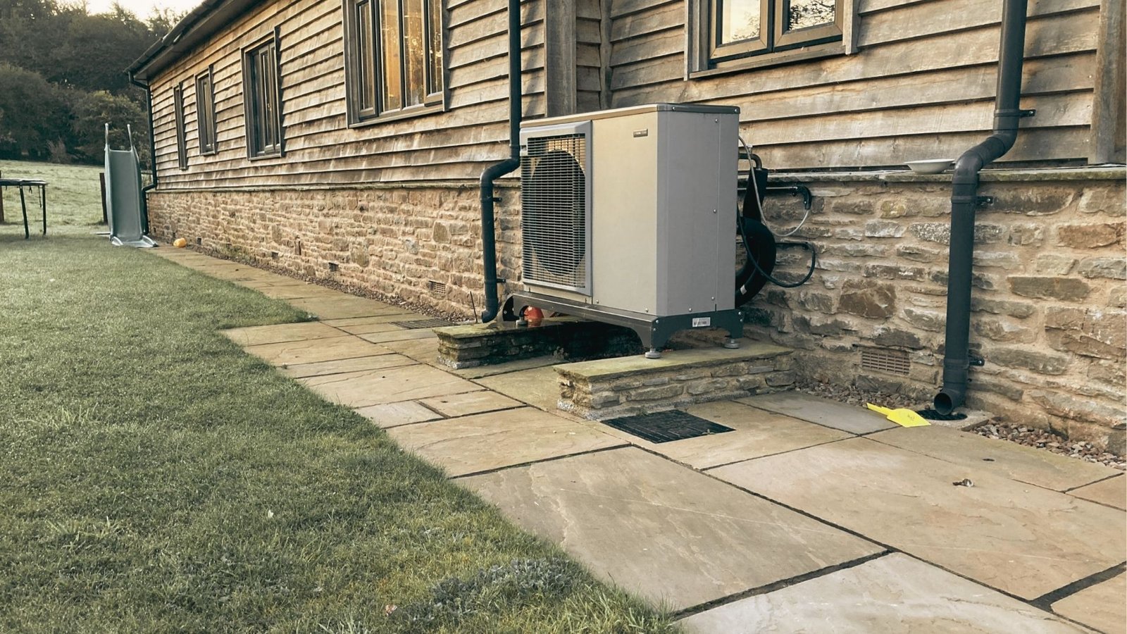 Outdoor heat pump unit mounted on a stone base beside a wooden country house.